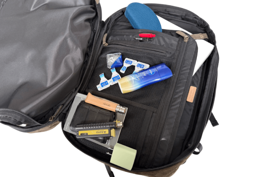 Backpack opened to reveal a toolkit: small bottles, tubes, a wallet, and various hardware items in built‑in pockets.
