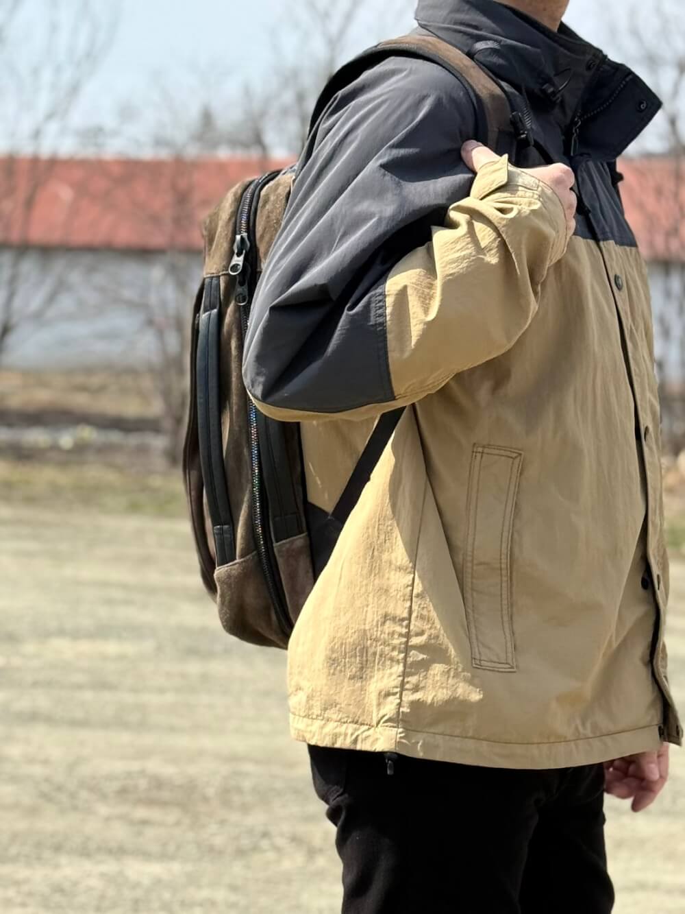 Person wearing a tan and black two-tone jacket with a brown backpack, outdoors, shown in profile from the side.