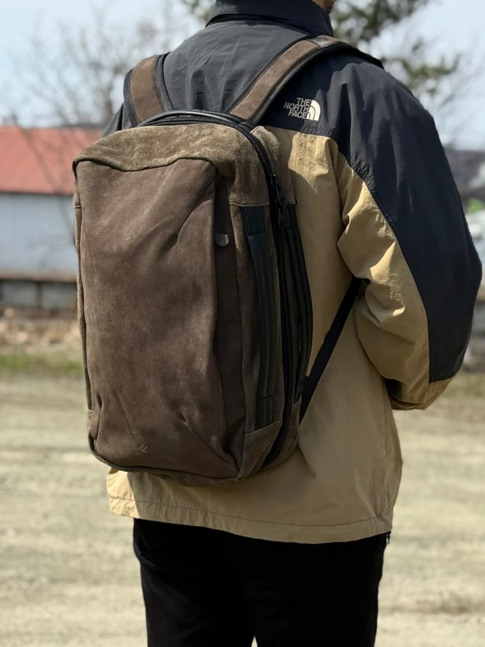 Person wearing a tan jacket with a brown backpack, viewed from behind outdoors.
