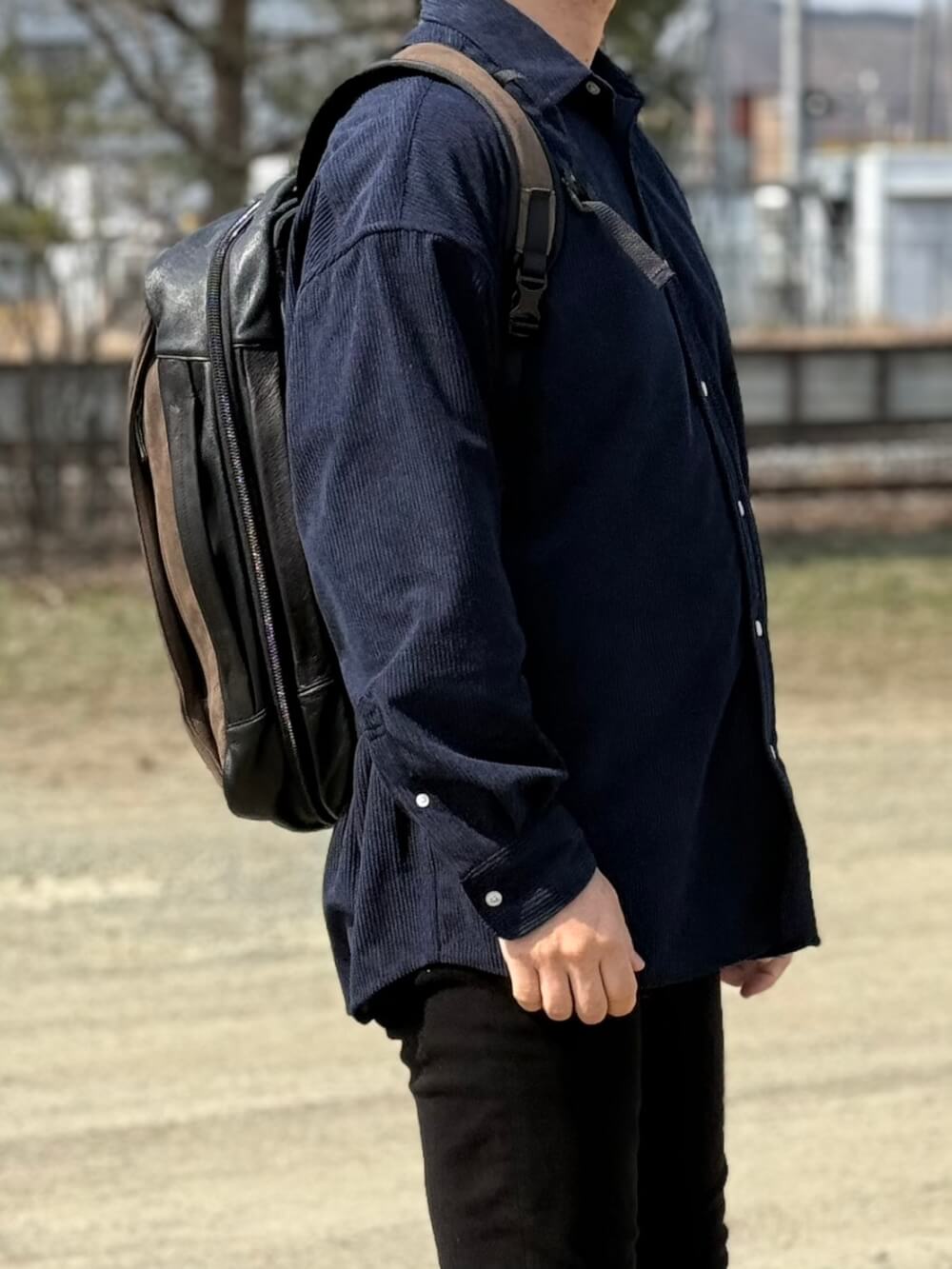 Person standing outdoors in a navy corduroy shirt with a black leather backpack on a daytime street or park setting.