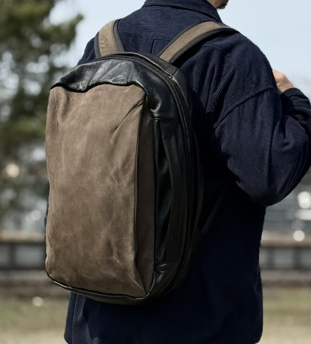 Person wearing a two-tone backpack (brown front, black sides) over a dark blue shirt, photographed from behind outdoors.