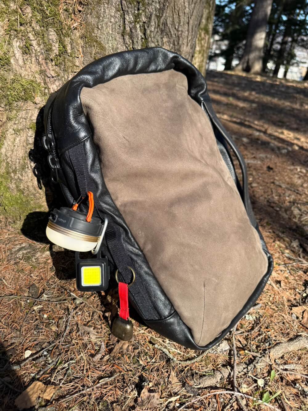 鹿革リュック 142 | 天然エゾ鹿革のレザレクション公式通販 | LEATHERECTION Backpack with a headlamp and small LED flashlight attached, resting against a tree outdoors.