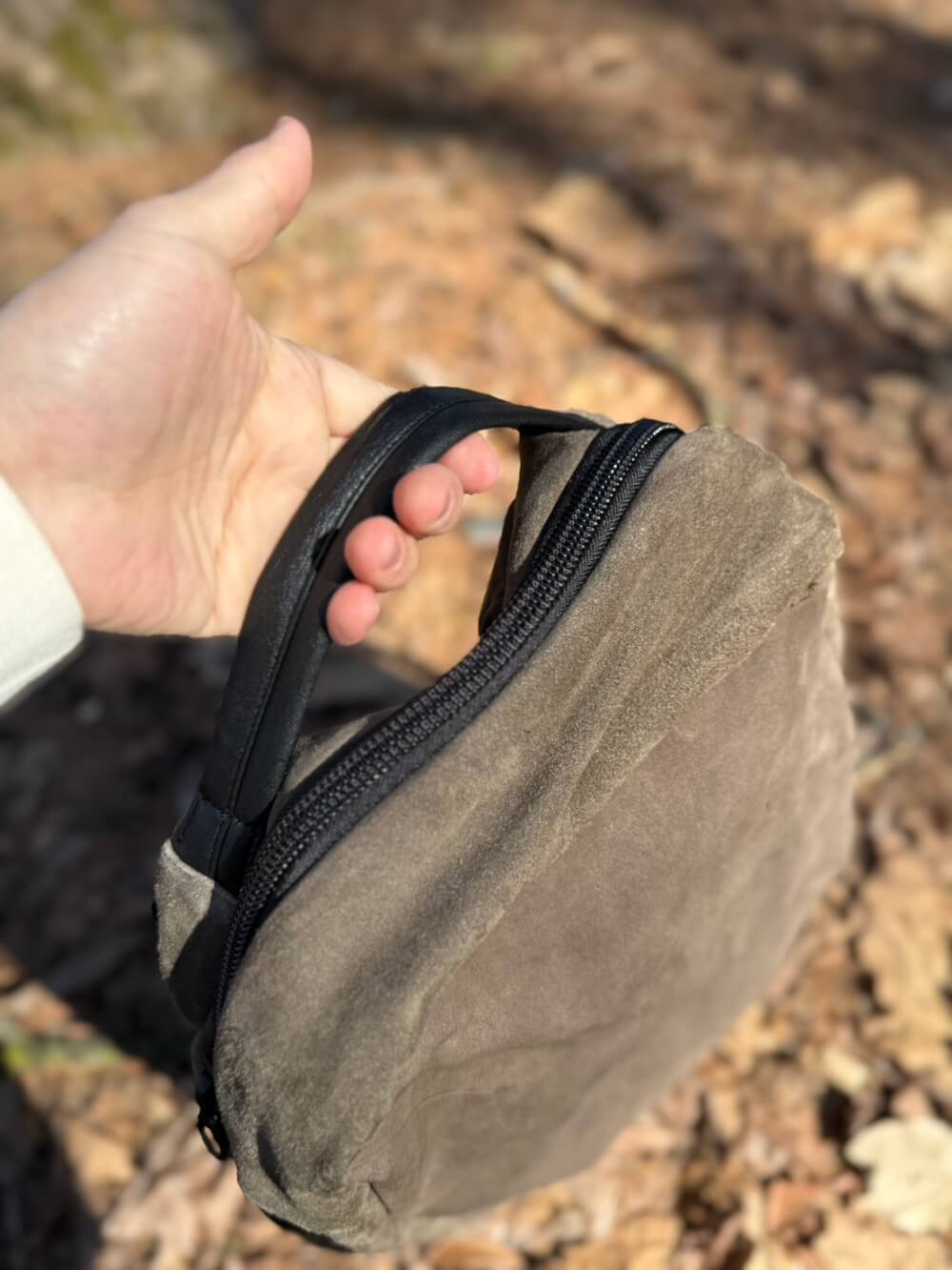 Hand holding a small brown suede zippered pouch outdoors on a leaf-strewn ground