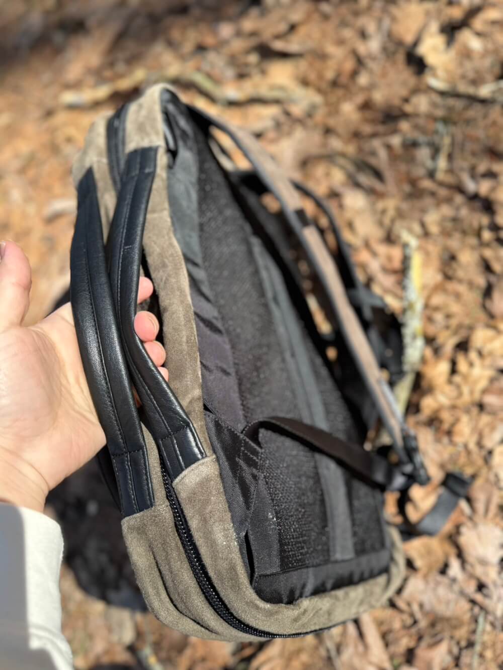 Hand holding a beige and black backpack open with zippers, outdoors on a leaf-covered ground