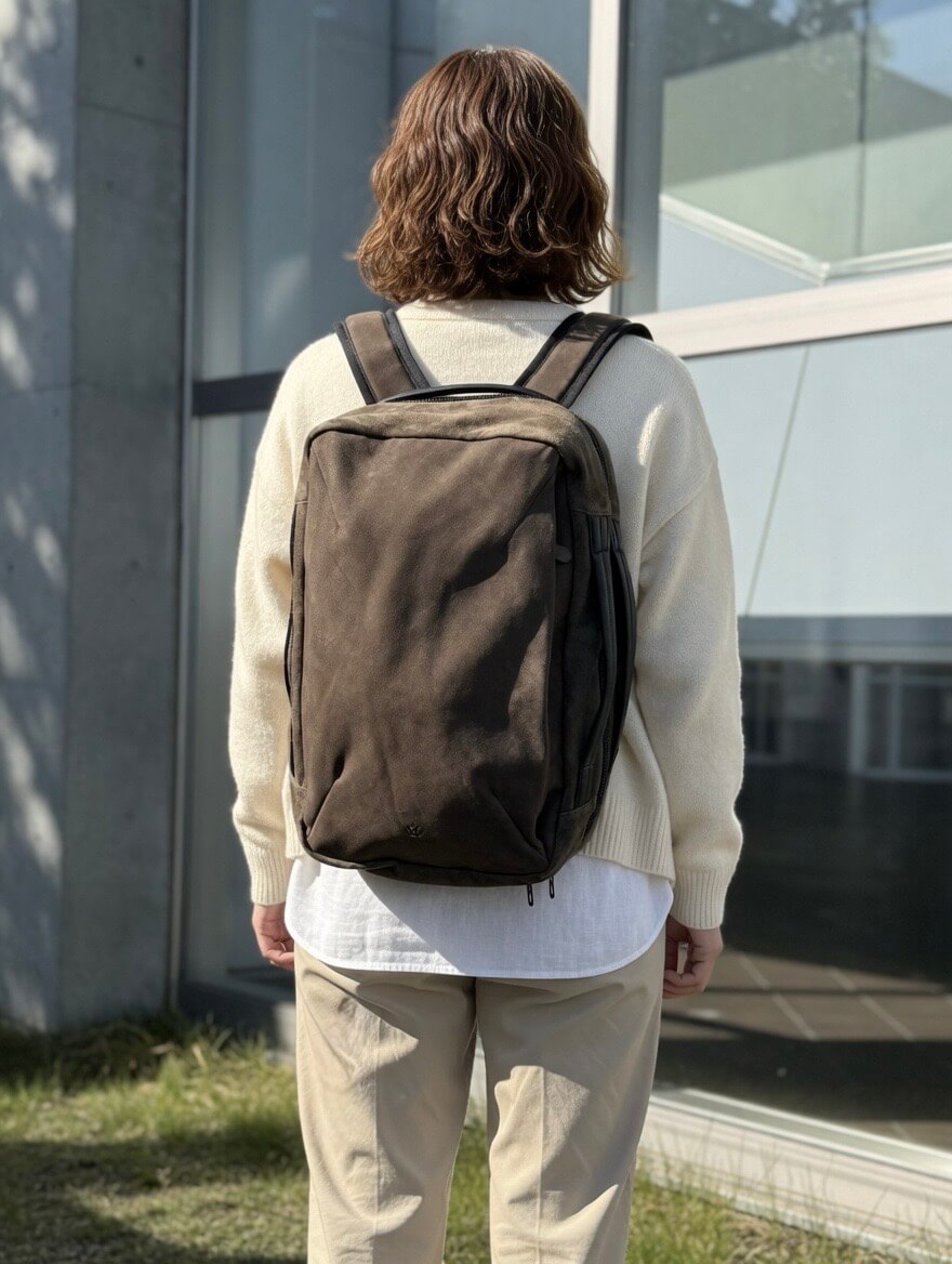 Person with shoulder-length brown hair wearing a cream sweater and white shirt, carrying a dark backpack outdoors near a glass building.