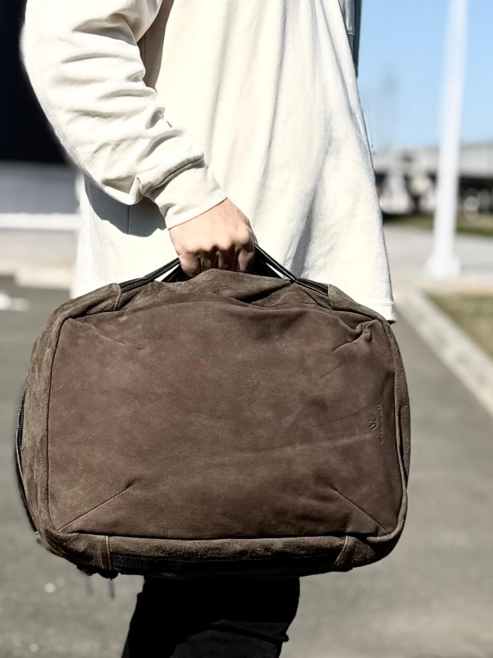 Person in a light beige top carrying a brown duffel bag by its handles while walking outdoors.