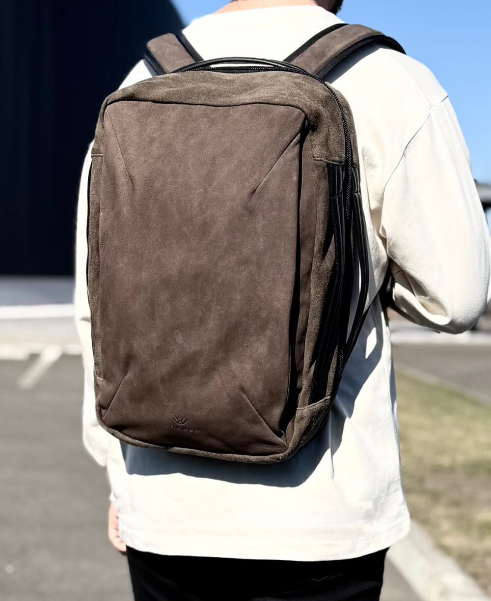 Person wearing a brown backpack viewed from behind outdoors. Back of a white jacketed figure in sunlight.