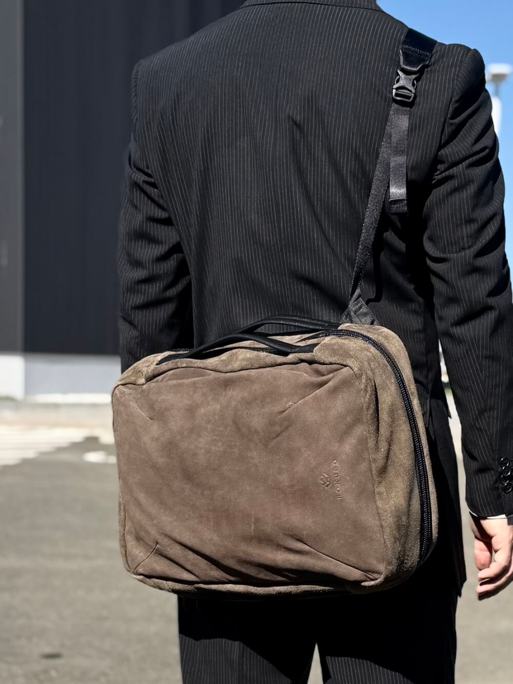 Person seen from behind wearing a black pinstripe suit and carrying a brown suede crossbody/messenger bag over the shoulder.