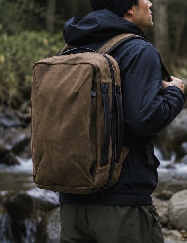 Person in a dark jacket and beanie wearing a brown backpack, standing by a rocky stream in a forested area.
