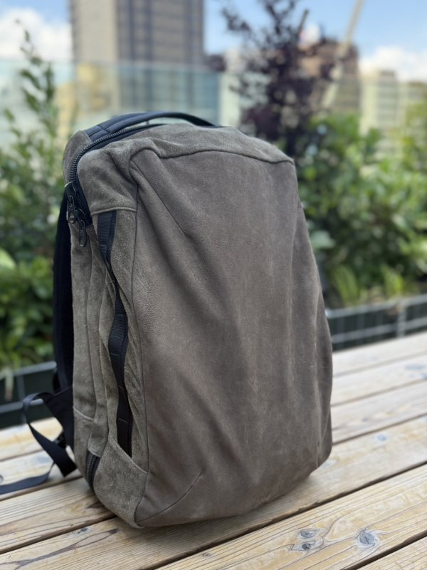 Gray backpack with black straps resting on a wooden outdoor surface, with greenery and buildings blurred in the background.