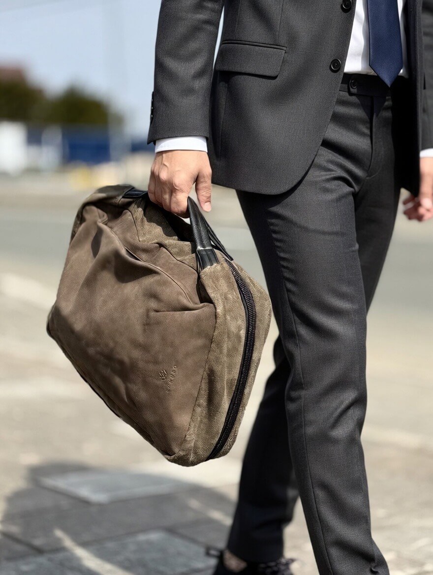 Businessperson in a dark suit carrying a brown duffel bag by its handle outdoors