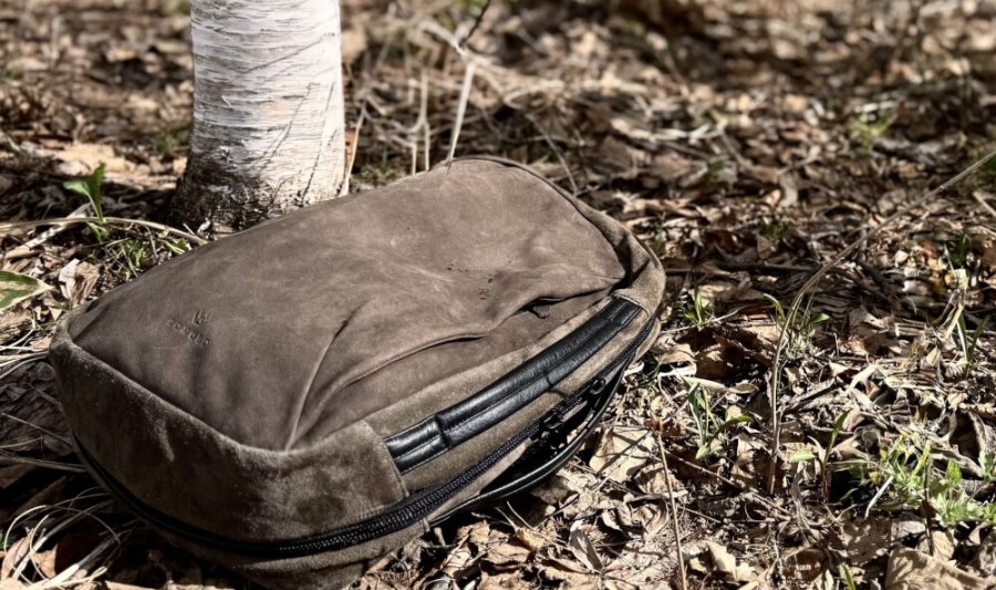 Brown padded camera bag lying on leaf-litter forest floor beside a tree trunk