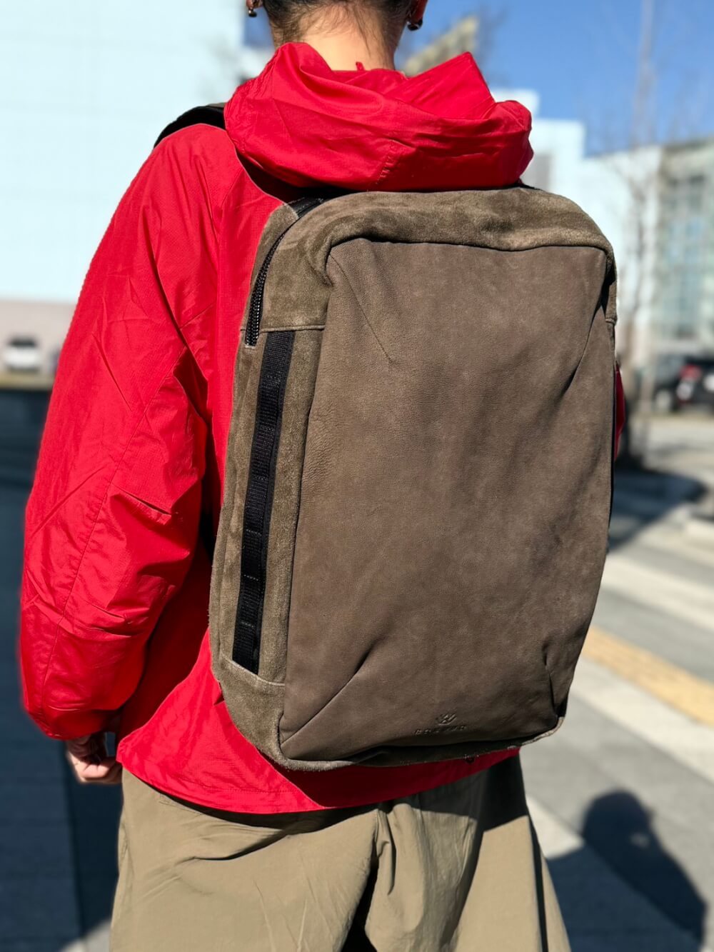 Person wearing a red jacket with a brown canvas backpack seen from the back on a sunny street