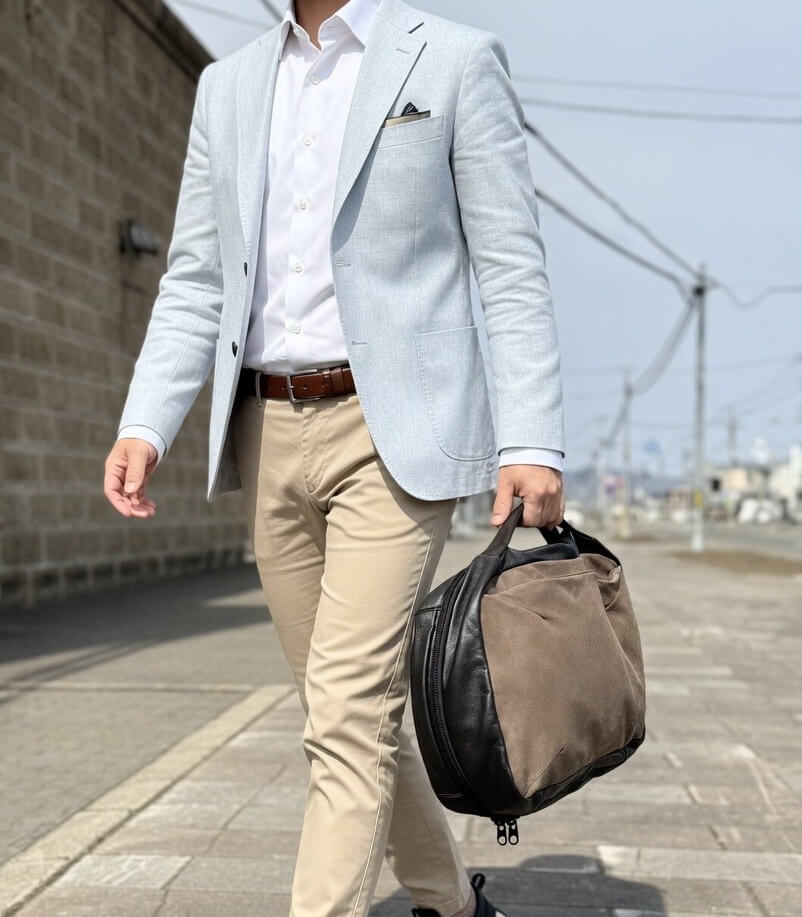 Man in a light gray blazer, white shirt, and beige pants walks along a sidewalk while carrying a brown-and-black duffel bag at his side.