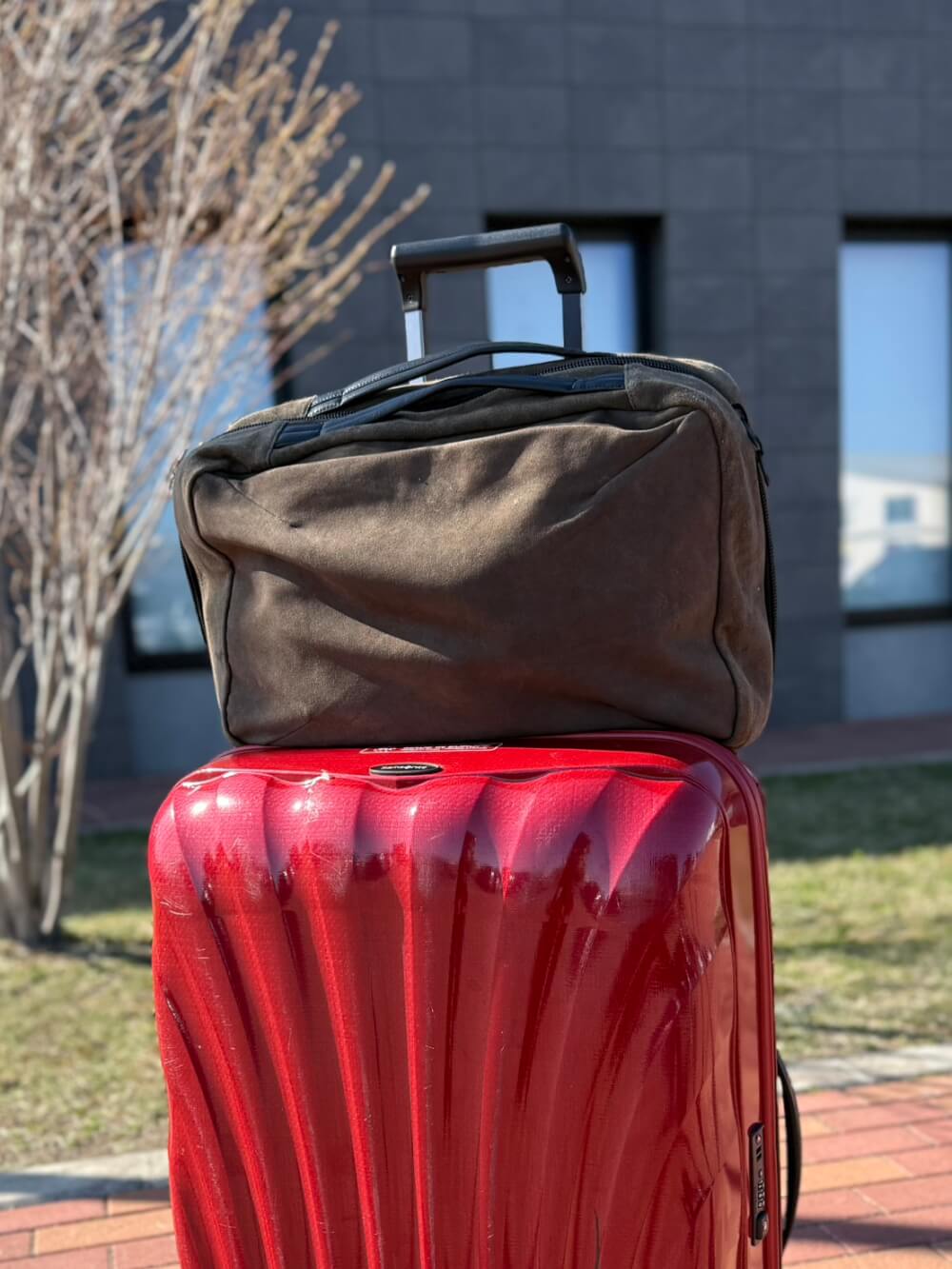Red hard-shell suitcase with a dark duffel bag on top, outdoors in front of a building.”