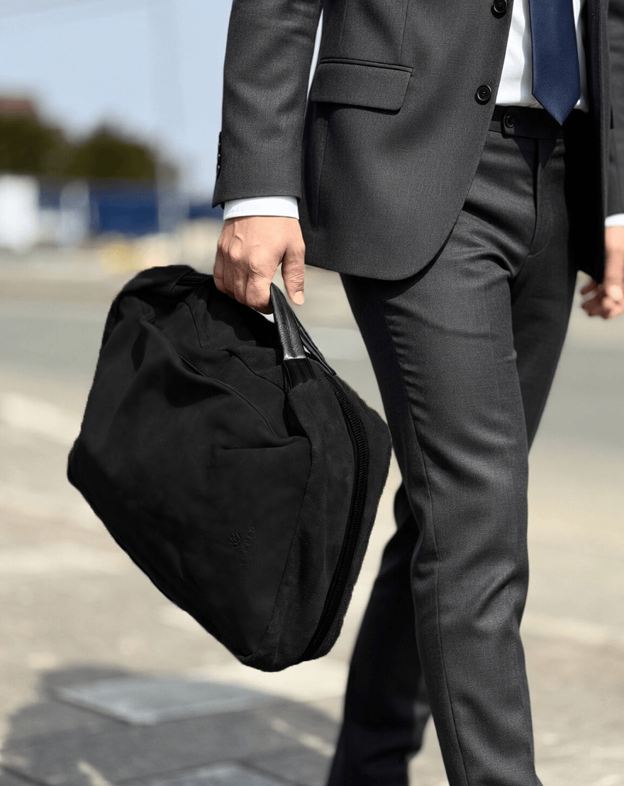 Businessperson in a gray suit walking outdoors while carrying a black duffel bag by its handle.