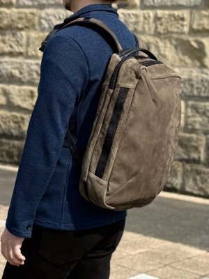 Person wearing a blue knit sweater carries a brown backpack outdoors by a stone wall.