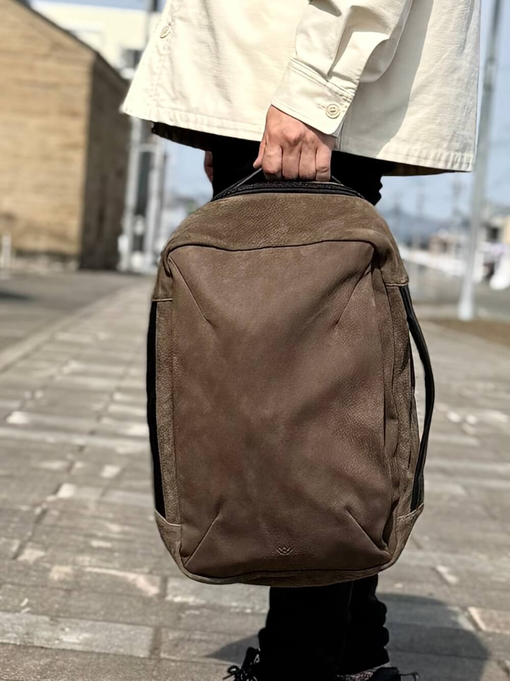 Person in a light jacket holding a brown backpack by the top handle on a sidewalk outdoor.