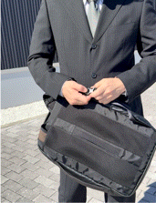 Man in a gray suit holding a black briefcase with a shoulder strap outdoors on a paved surface.
