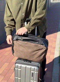 Person in an olive-green shirt grips a brown shoulder bag placed on a silver wheeled suitcase on a brick pavement.