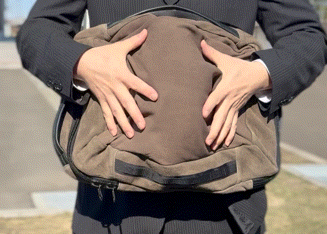 Person in a dark suit holding a brown duffel bag in front of their torso, gripping the handles.